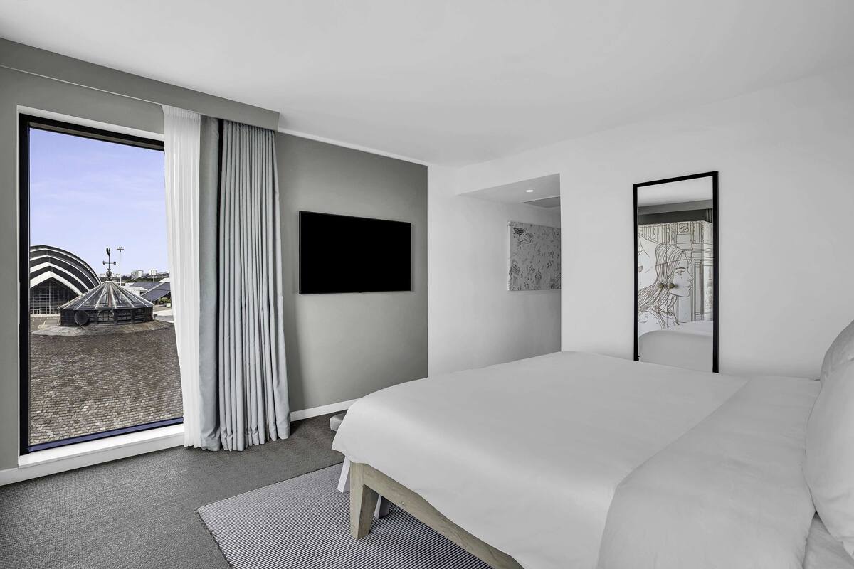 Ein modernes Hotelzimmer mit einem großen Bett, einem Flachbildfernseher, einem großen Fenster mit Vorhängen und Blick auf einen Außenbereich.