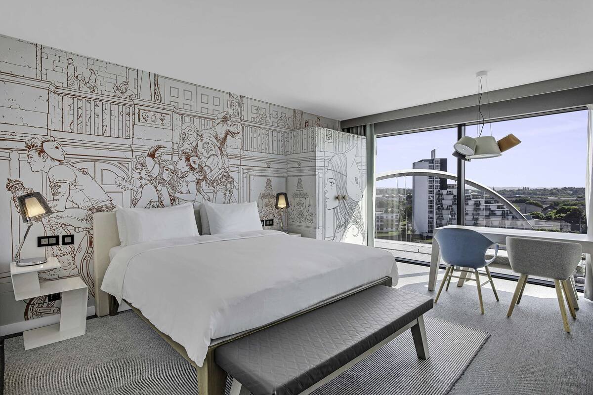Ein modernes Hotelzimmer mit einem großen Bett, einem Schreibtisch mit zwei Stühlen, einem großen Fenster mit Blick auf die Stadt und einer Wanddekoration mit Weltkartenmuster.