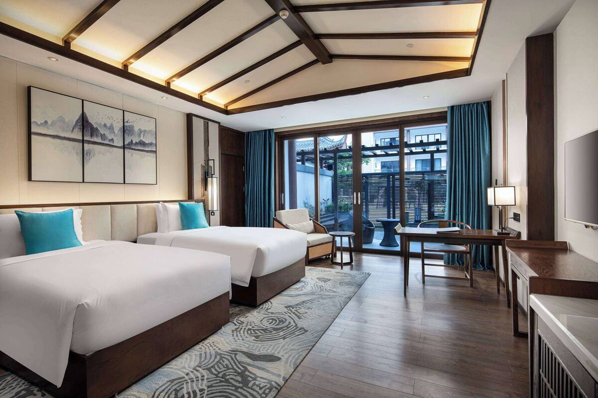 A hotel room with two beds, a desk, a chair, and a large window with a view of a traditional building.