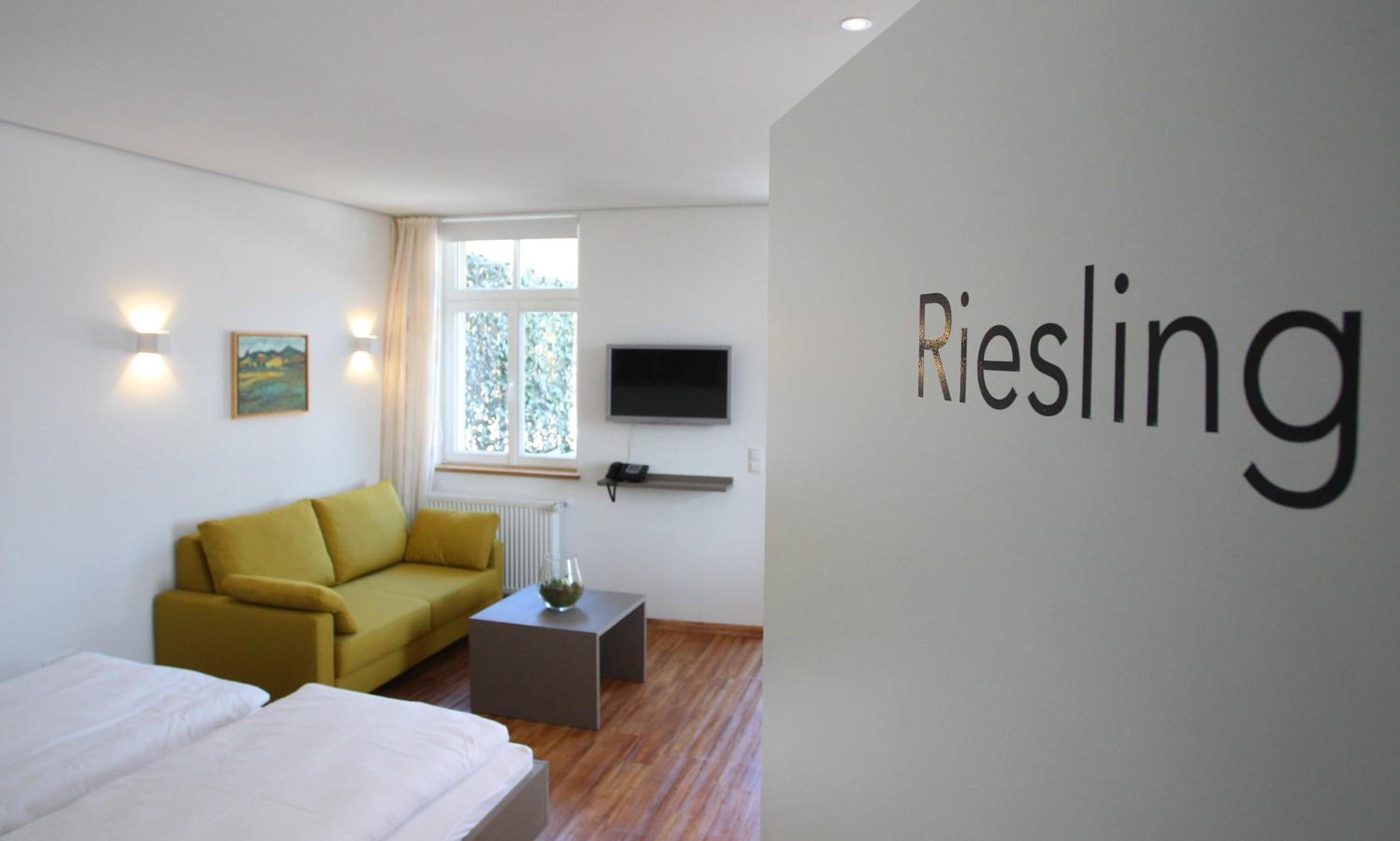 A modern hotel room with a yellow sofa, a bed, a small table with a vase, a TV mounted on the wall, and a window with a view of trees.
