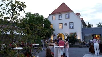 An outdoor courtyard with a building featuring a red-tiled roof and multiple windows. There are several round tables and chairs arranged for outdoor dining.