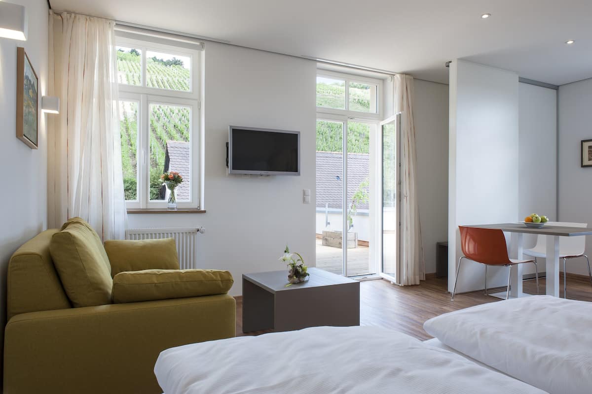A modern hotel room with a bed, a sofa, a small table with a vase, a TV, and a dining area with a chair and table.
