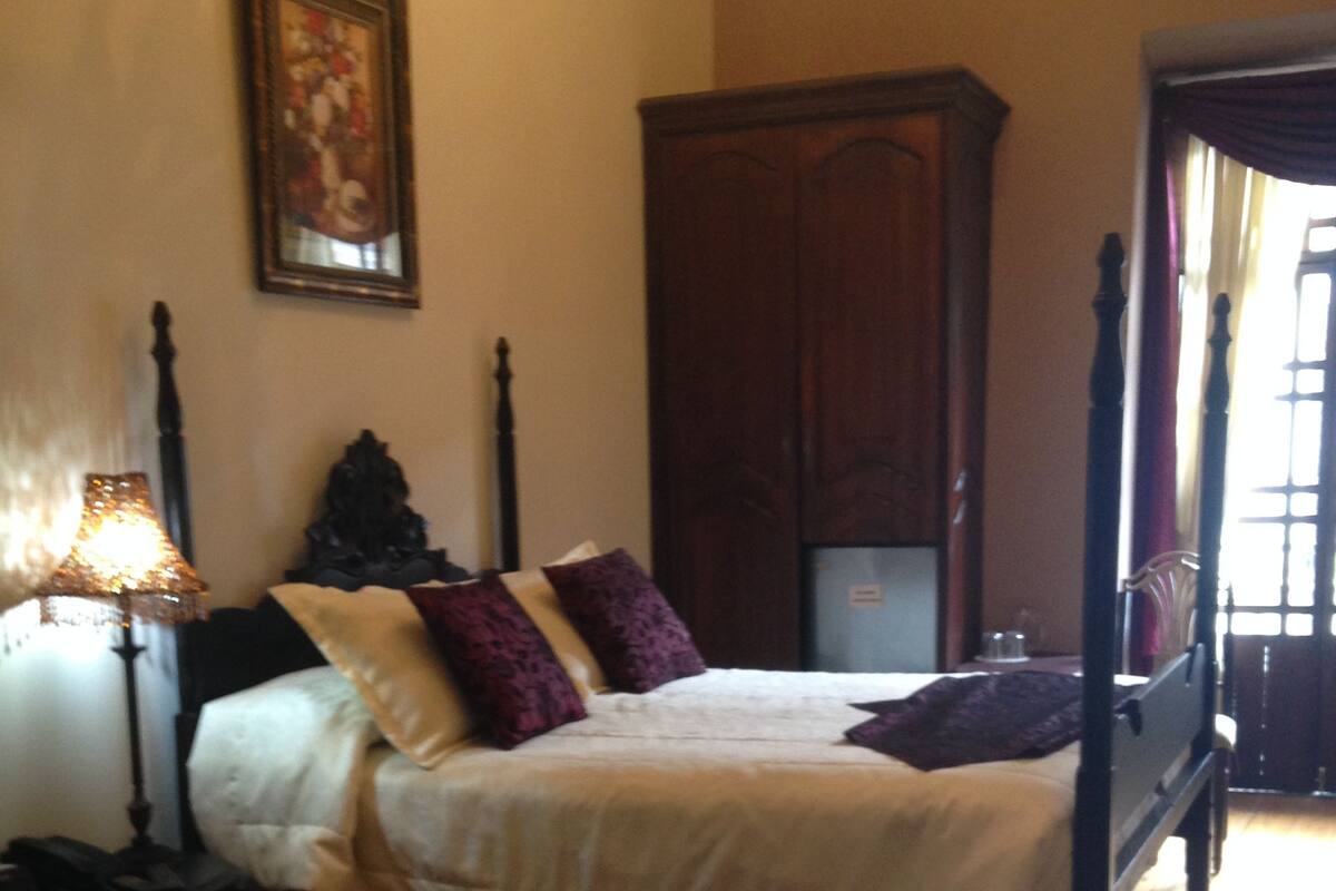 Un dormitorio con una cama con dosel, una mesita de noche con lámpara, un armario de madera y un cuadro enmarcado en la pared.