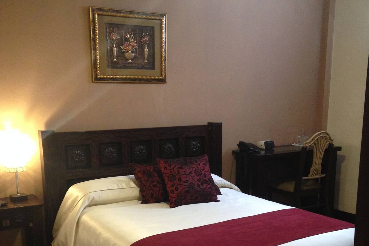 Una cama bien hecha con una manta roja, un cabecero de madera y un cuadro en la pared.