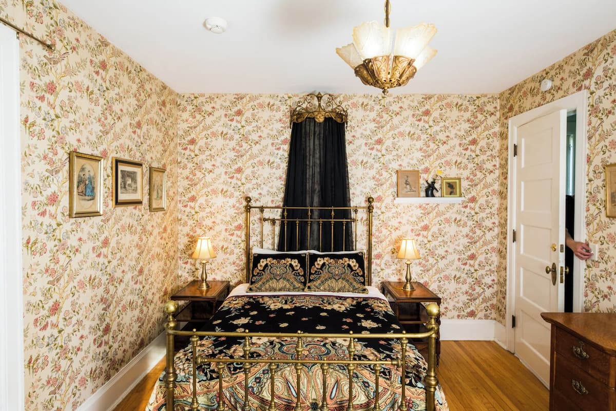Ein Schlafzimmer mit floralem Wandpapier, einem Bett mit gemustertem Überwurf, zwei Nachttischlampen, einer Kandelaber und einer Tür.