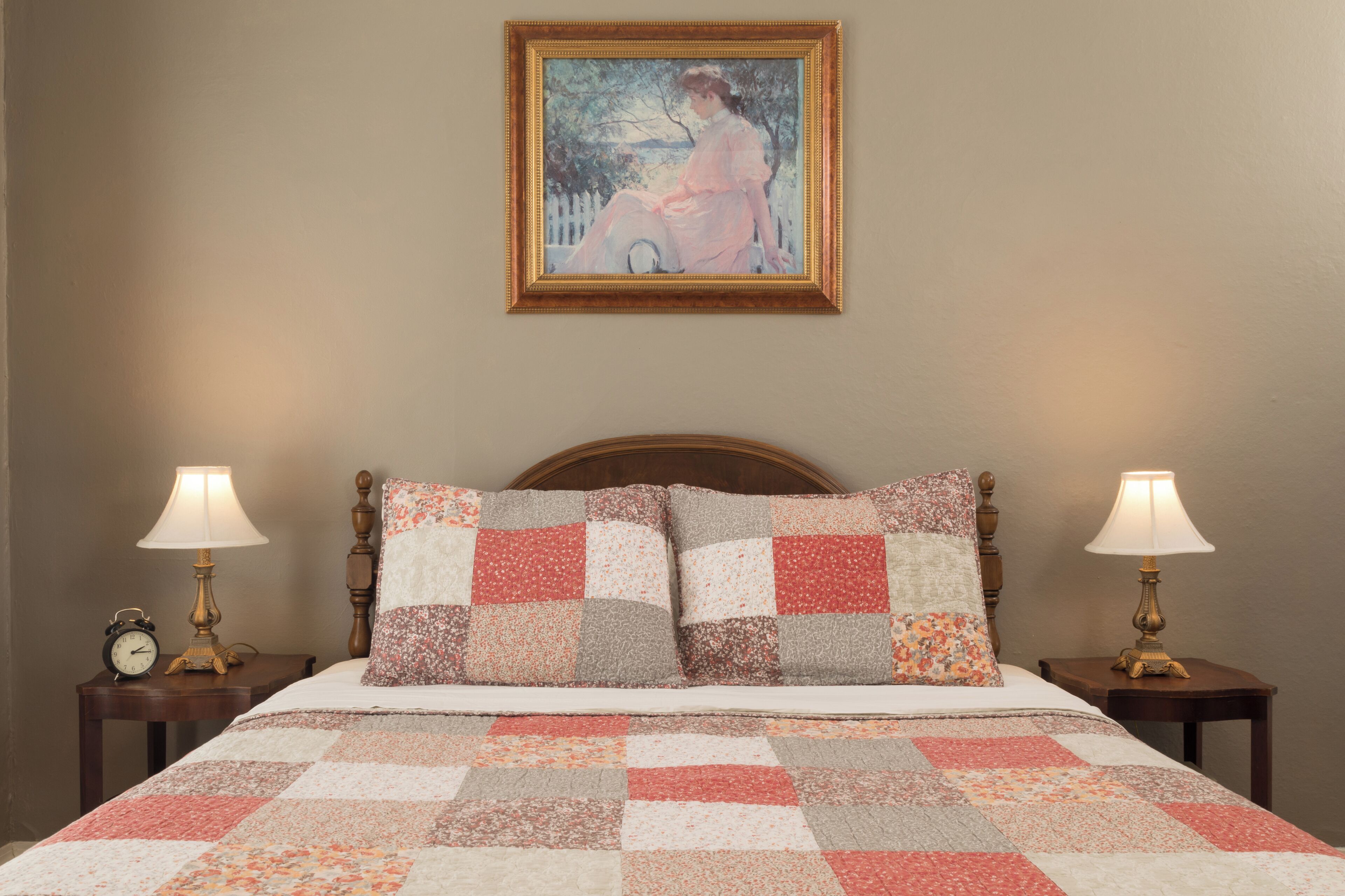A bedroom with a wooden headboard, a quilted bedspread, two bedside lamps, and a framed painting on the wall.