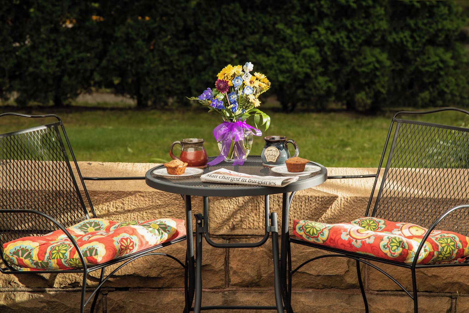 Un espacio al aire libre con una mesa y sillas de metal, un jarrón de flores, una taza, una tetera y un muffin sobre un periódico.