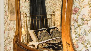 A wooden dresser with a mirror, a lamp, and a bed with floral wallpaper.