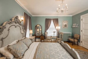 Ein Schlafzimmer mit einem großen Bett, einer Kristallleuchter, einem Schminktisch, einem Sessel und einem Fenster mit Vorhängen.