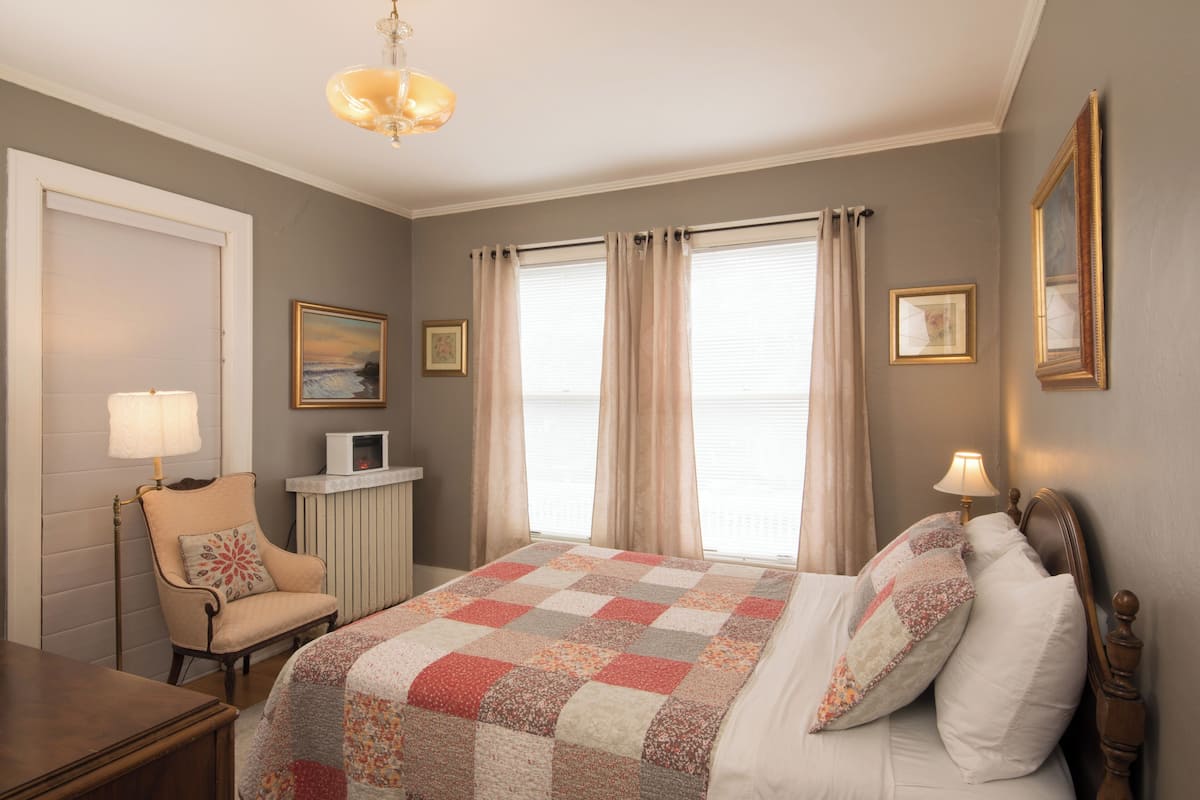 Ein Schlafzimmer mit einem Bett, einem Sessel, einem Schreibtisch, einer Lampe, einem Bilderrahmen und einem Fenster mit Vorhängen.