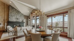 A spacious dining room with a large round table, comfortable seating, a stone fireplace, and large windows offering a view of the landscape.