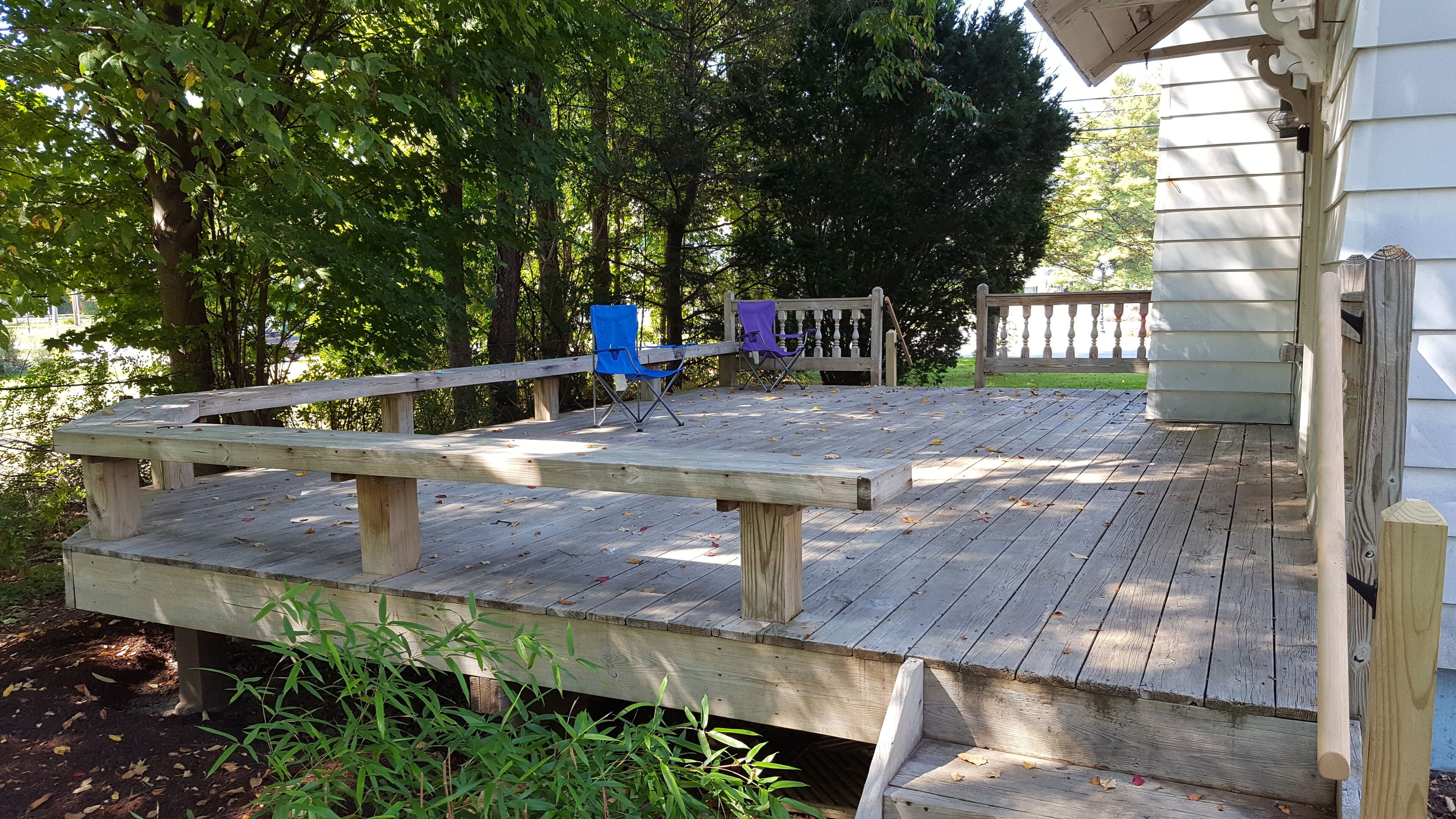 A wooden deck with a ramp leading to a covered porch area, surrounded by trees and a fence.