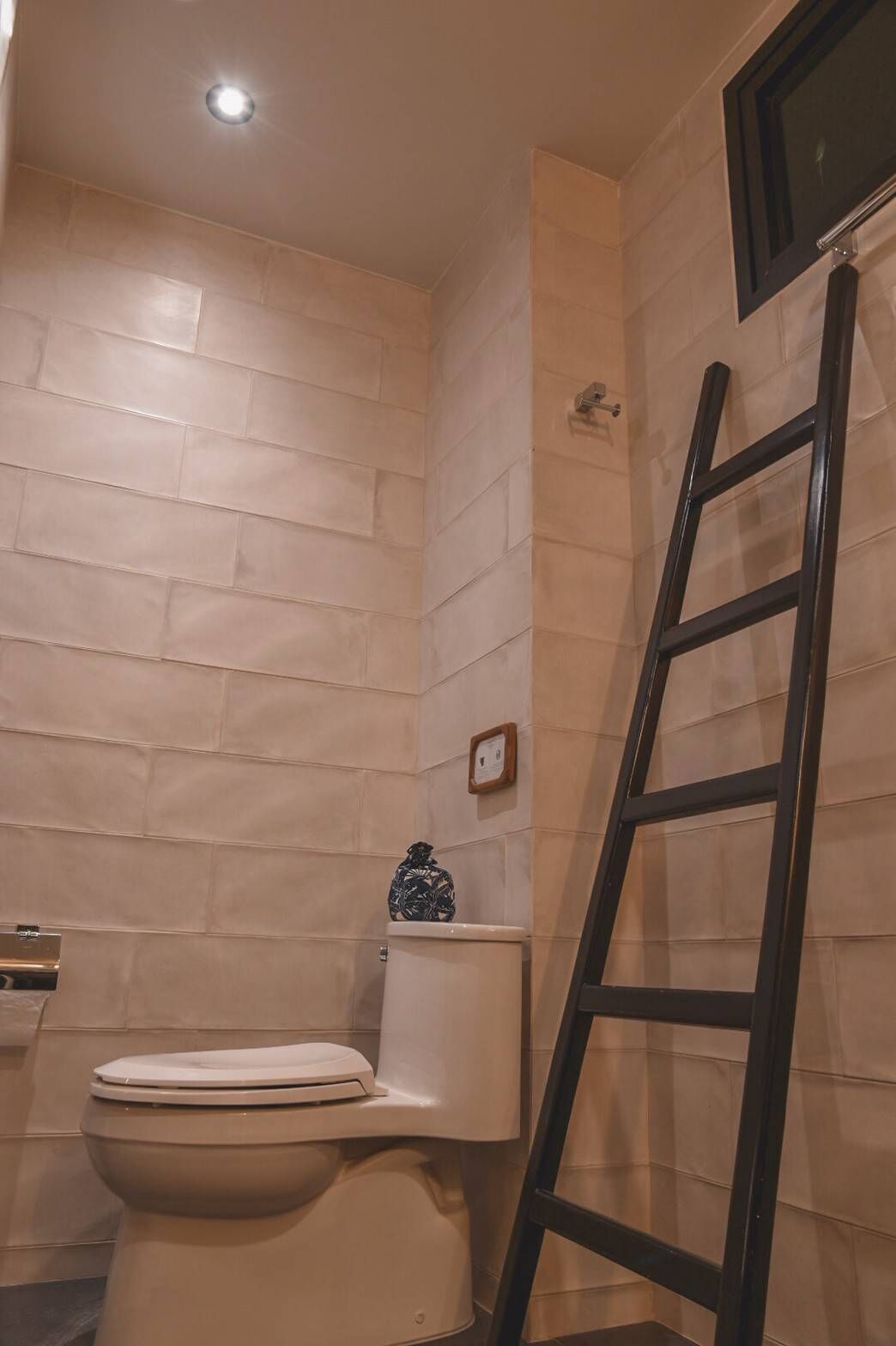 A bathroom with beige tiled walls, a white toilet, a black metal ladder, and a small framed picture on the wall.