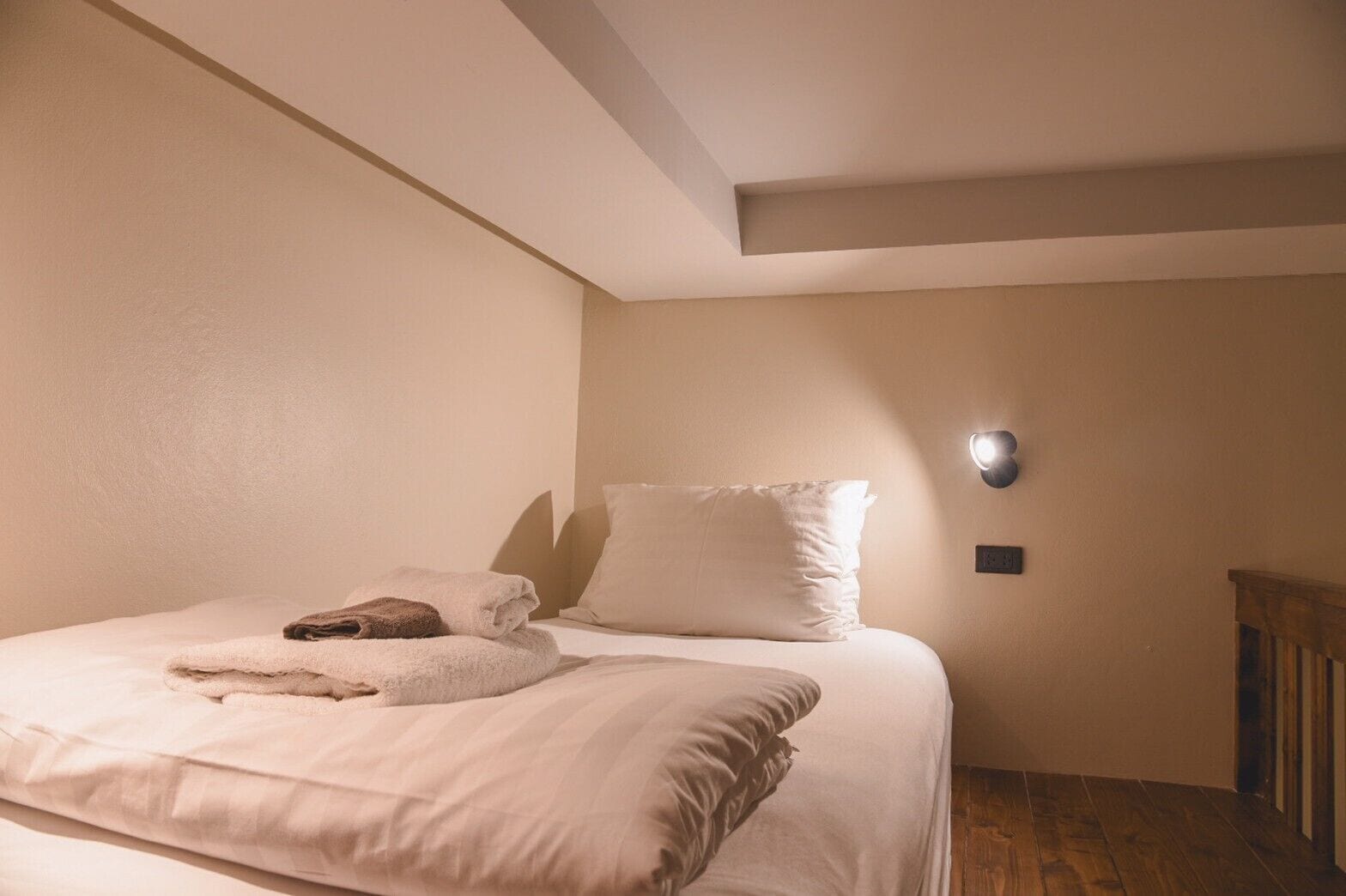 Una cama bien hecha con una toalla doblada y una almohada, un aplique de pared y suelo de madera.