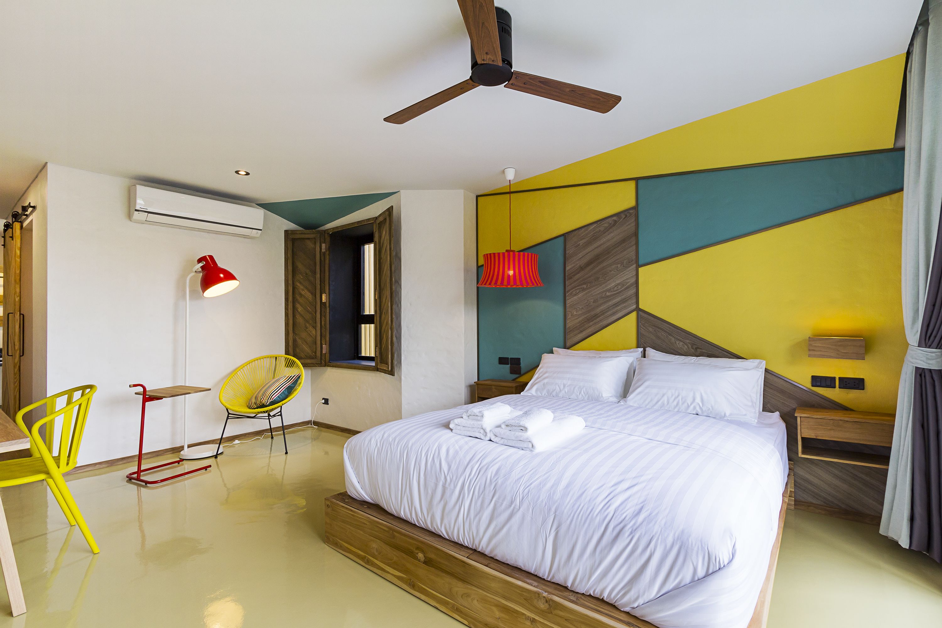 A modern bedroom with a bed, a ceiling fan, a red lamp, a yellow chair, and a wooden nightstand.