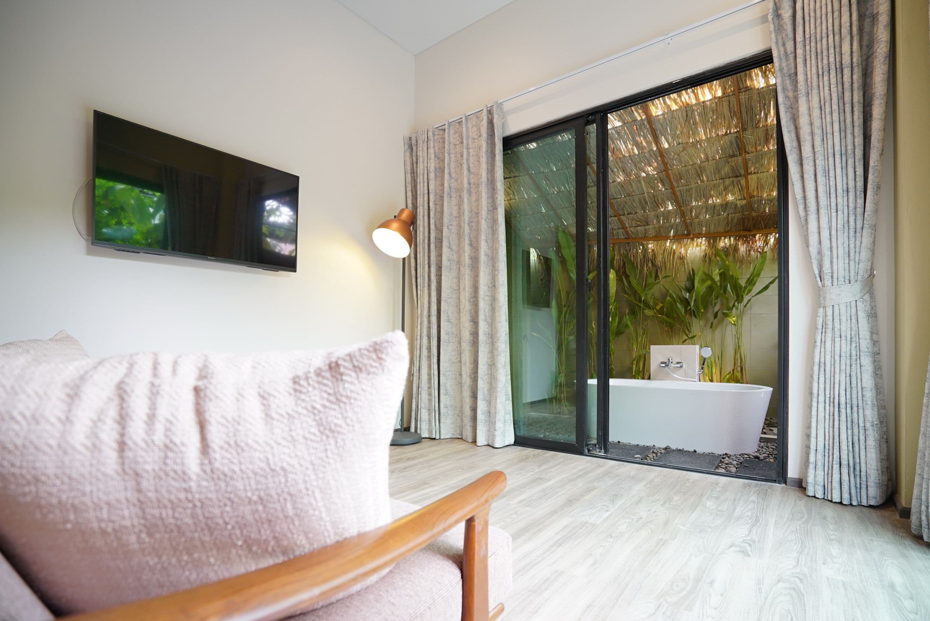 A modern living room with a flat-screen TV, a wooden chair, a large window with curtains, and a view of a garden and outdoor area.