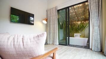 Un salon moderne doté d’un téléviseur à écran plat, d’un fauteuil en bois, d’une grande fenêtre avec des rideaux et offrant une vue sur un jardin et un espace extérieur.