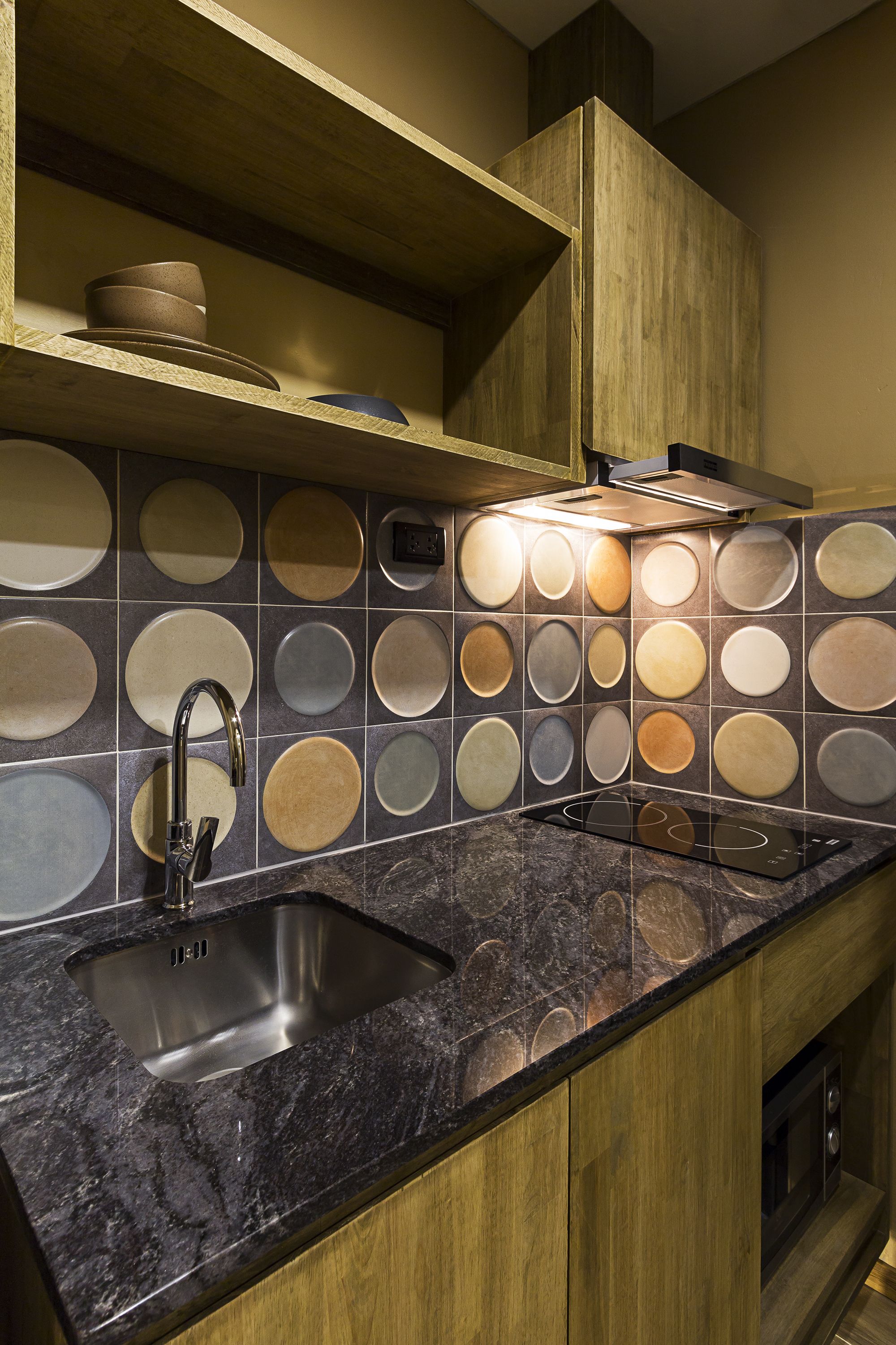 A kitchen with a granite countertop, stainless steel sink, and a wall with circular tiles.
