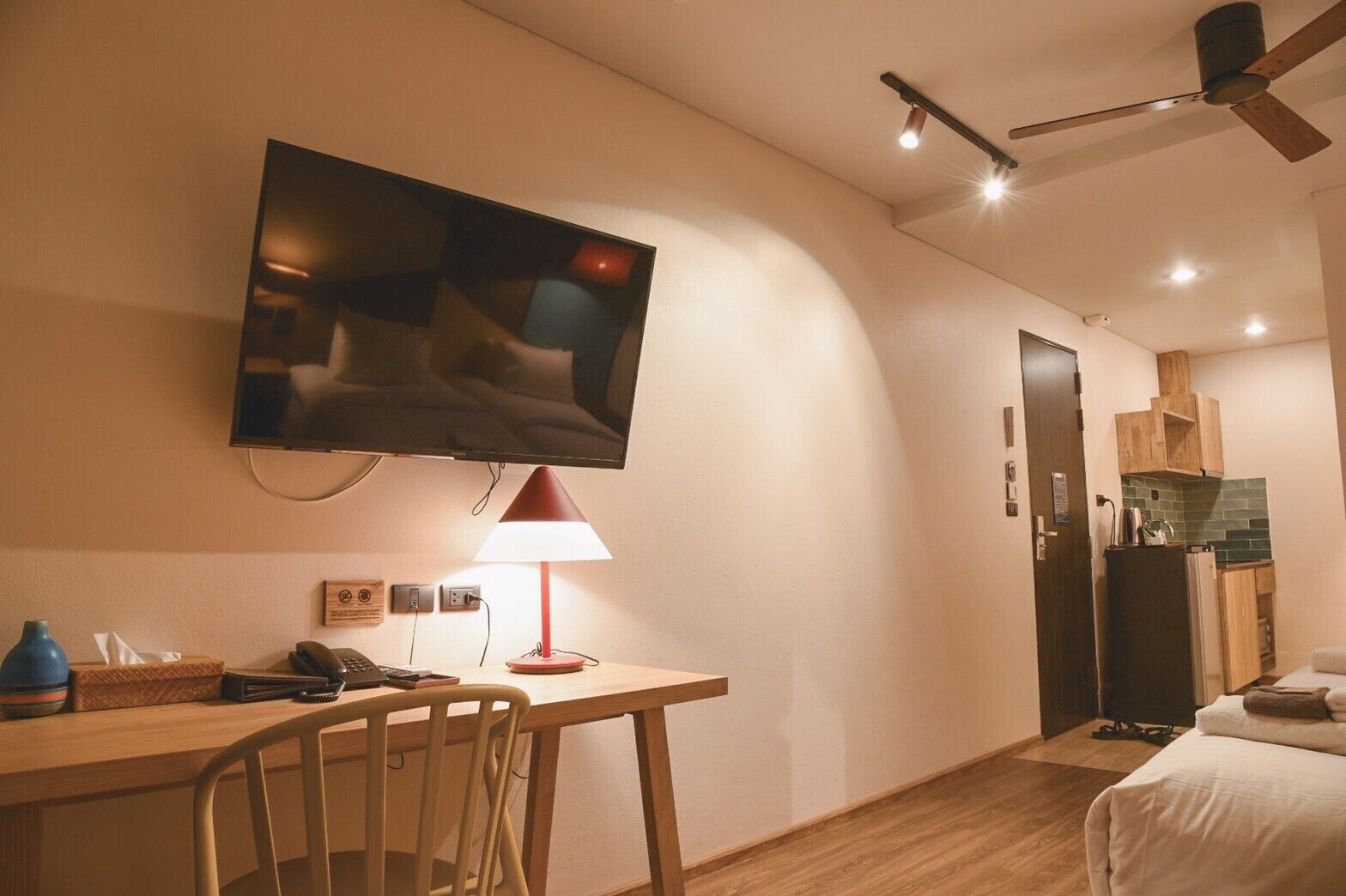 Habitación de hotel con televisor de pantalla plana en la pared, un escritorio con lámpara, una silla, una cama con edredón blanco y un área de kitchenette.