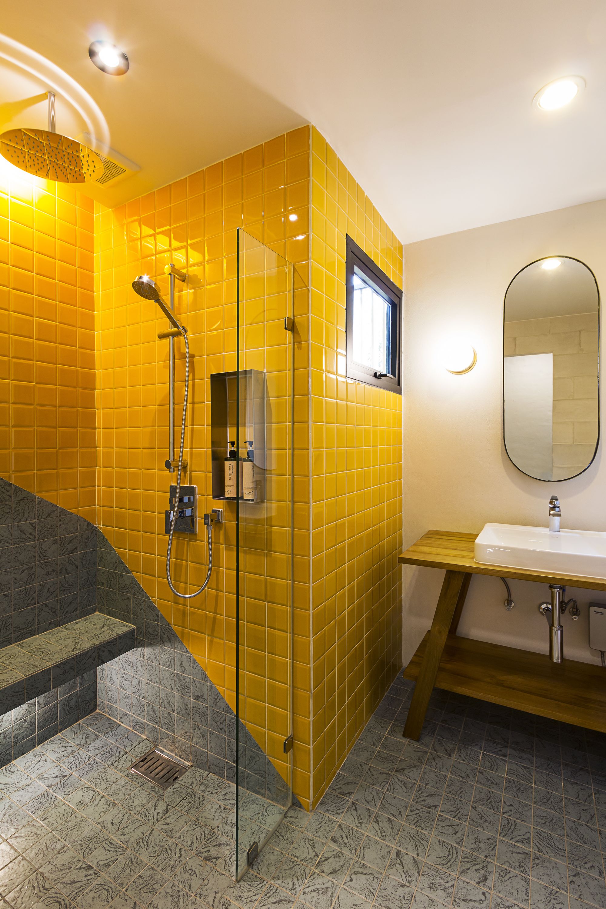 A bathroom with yellow tiled walls, a glass shower enclosure, a wooden vanity, and a marble-patterned floor.