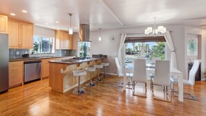 Une cuisine moderne avec des armoires en bois, un réfrigérateur en acier inoxydable, un îlot central intégré avec des tabourets de bar et une grande fenêtre offrant une vue sur l’extérieur.