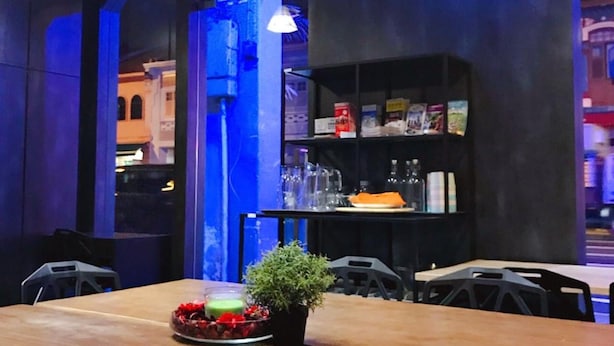 En restaurant med et træbord, stole, en lille plante og en reol med glas og bøger.