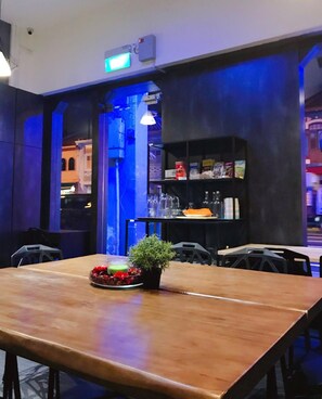 A restaurant with a wooden table, chairs, a small plant, and a shelf with glasses and books. - Atlantis Pods at Little India (Singapore)