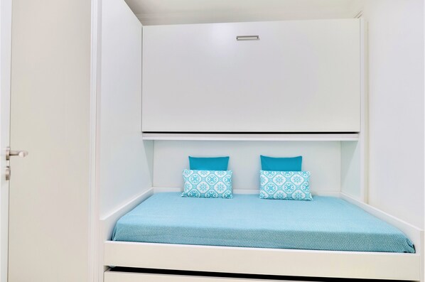 A compact room with a bed, two pillows, and a small overhead shelf. - Vale do Lobo Apartment B (Vale do Lobo)