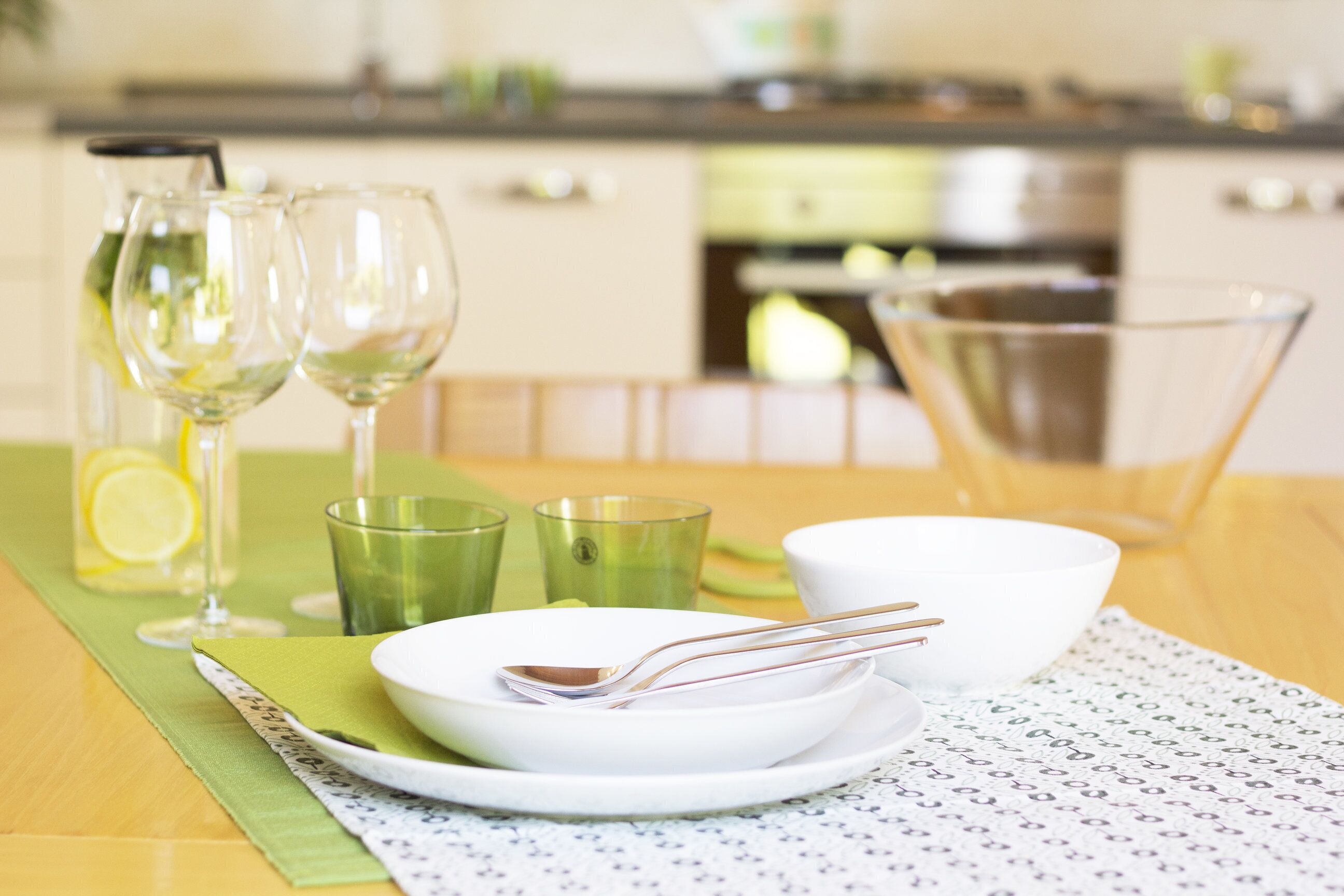 A dining table with a green placemat, a white plate with a spoon, a white bowl, and a green glass.