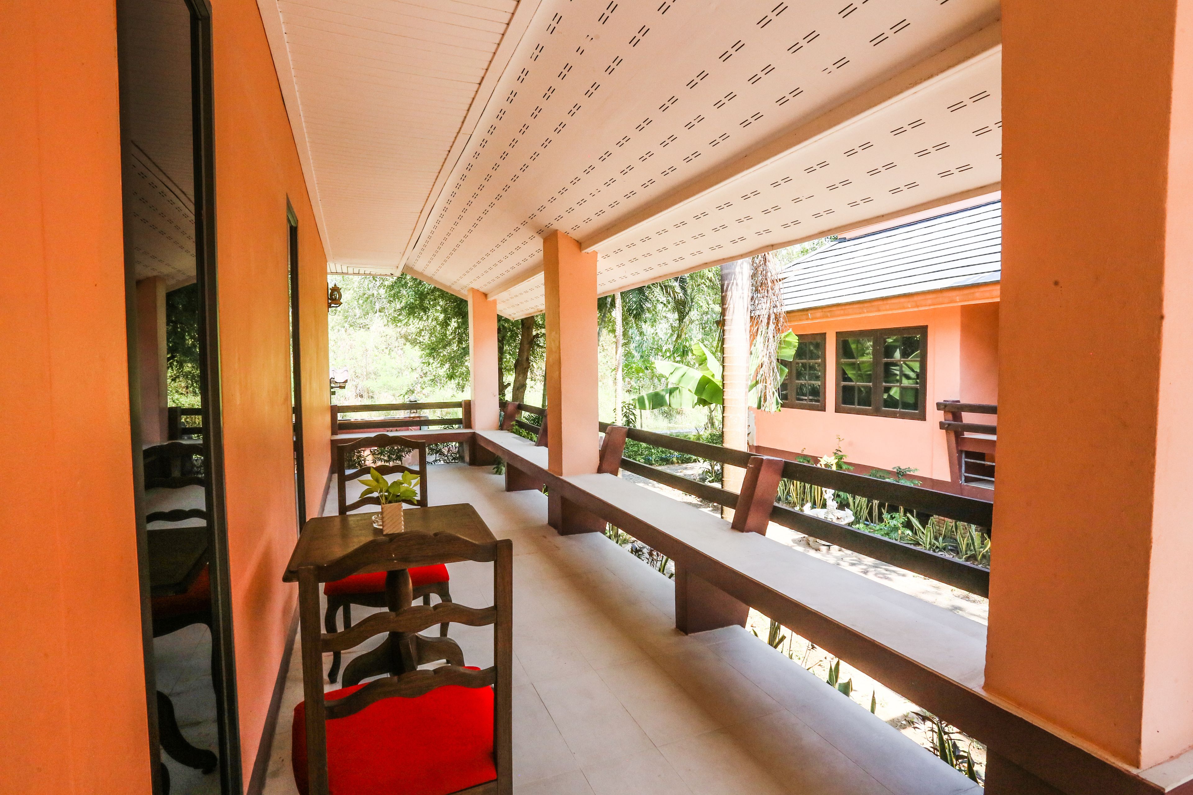 Un área de estar al aire libre con techo, con bancos de madera y sillas rojas, con vista a un jardín y un edificio de fachada rosa.