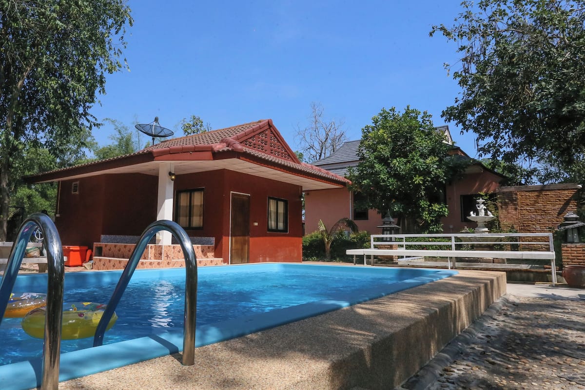 Una piscina con un edificio de techo rojo al fondo, rodeada de árboles y una valla blanca.