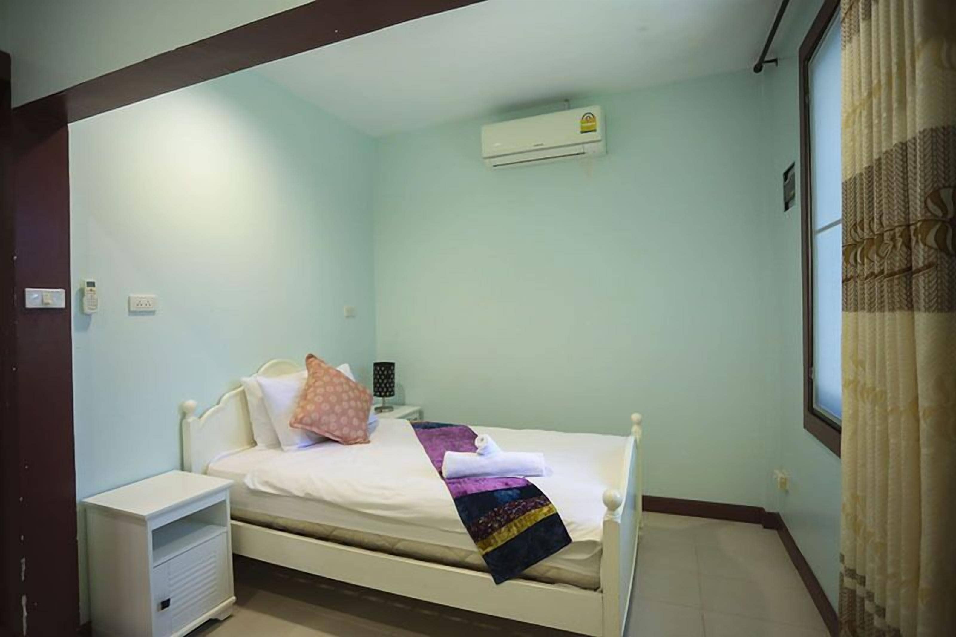 Habitación individual con una cama blanca, una mesita de noche, un equipo de aire acondicionado y una ventana con cortinas.