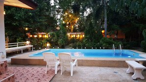 Ein Swimmingpool mit zwei weißen Plastikstühlen, umgeben von üppiger Vegetation und dekorativer Beleuchtung.