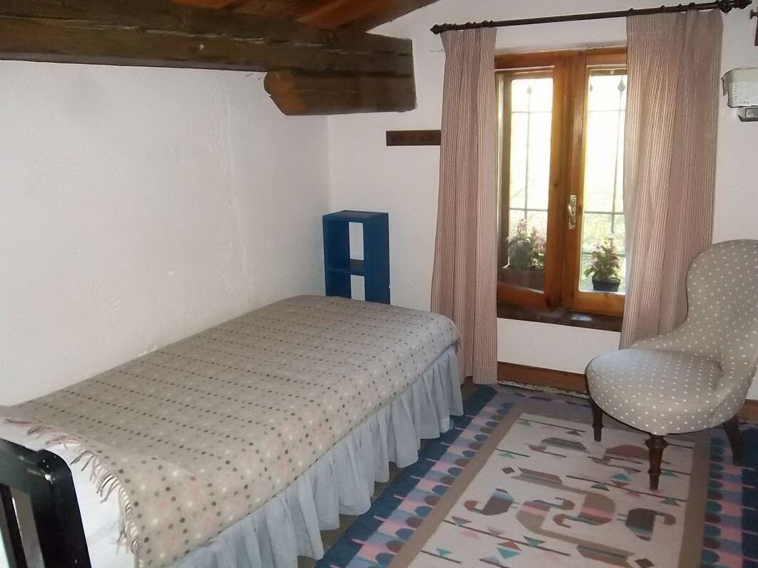 A small, single-bed room with a wooden ceiling, a patterned bedspread, a blue stool, a polka-dotted chair, and a window with curtains.