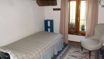 A small, single-bed room with a wooden ceiling, a patterned bedspread, a blue stool, a polka-dotted chair, and a window with curtains.