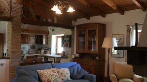 A cozy living room with a blue sofa, a beige armchair, and a fireplace. A kitchen with wooden cabinets and a dining area is visible in the background.