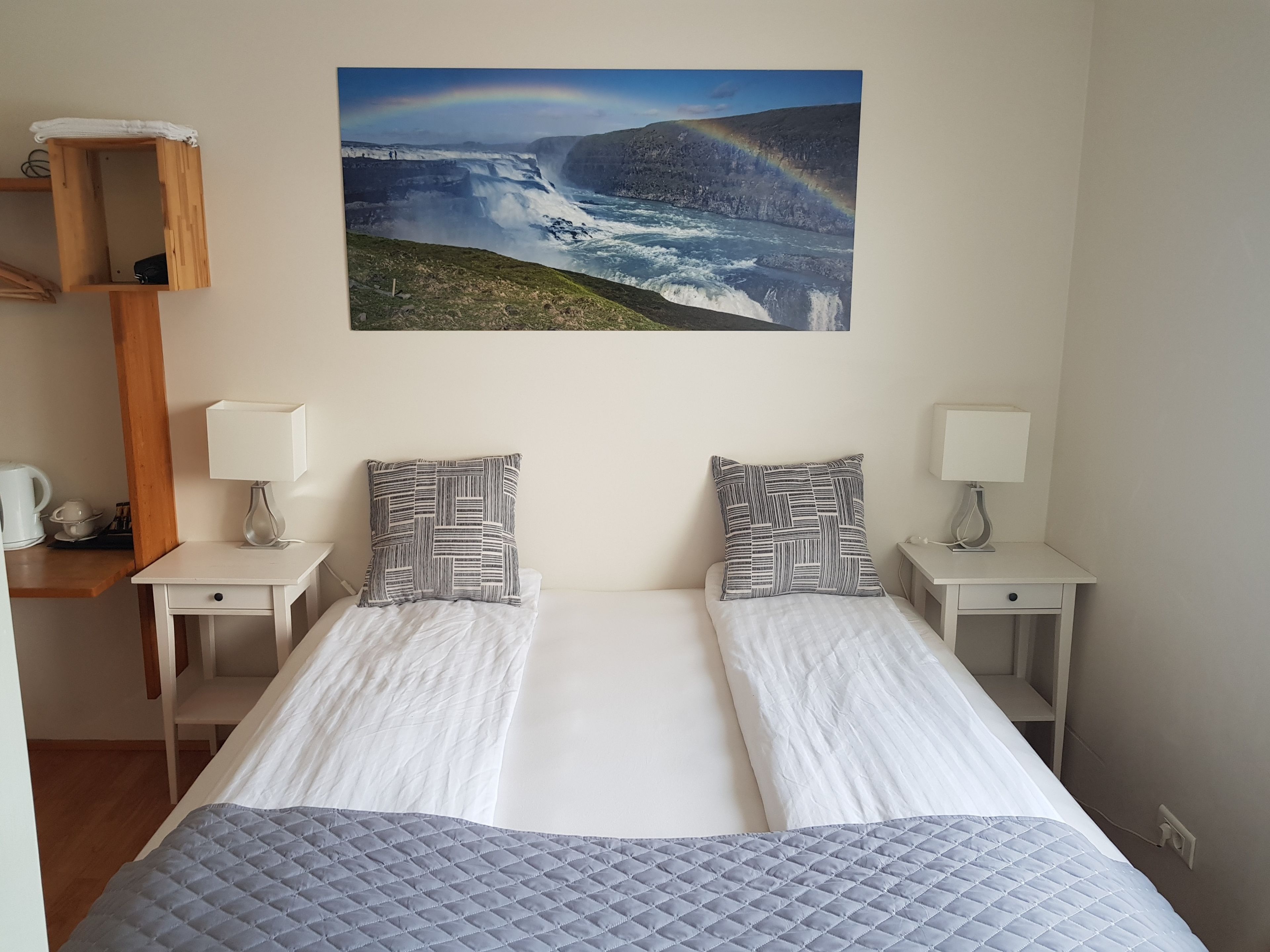 A double bed with a quilted grey bedspread, two bedside tables with lamps, a wooden shelf, and a framed picture of a waterfall with a rainbow.