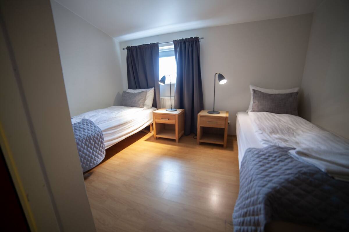 A small, well-lit bedroom with two single beds, a nightstand with a lamp, and a window with curtains.