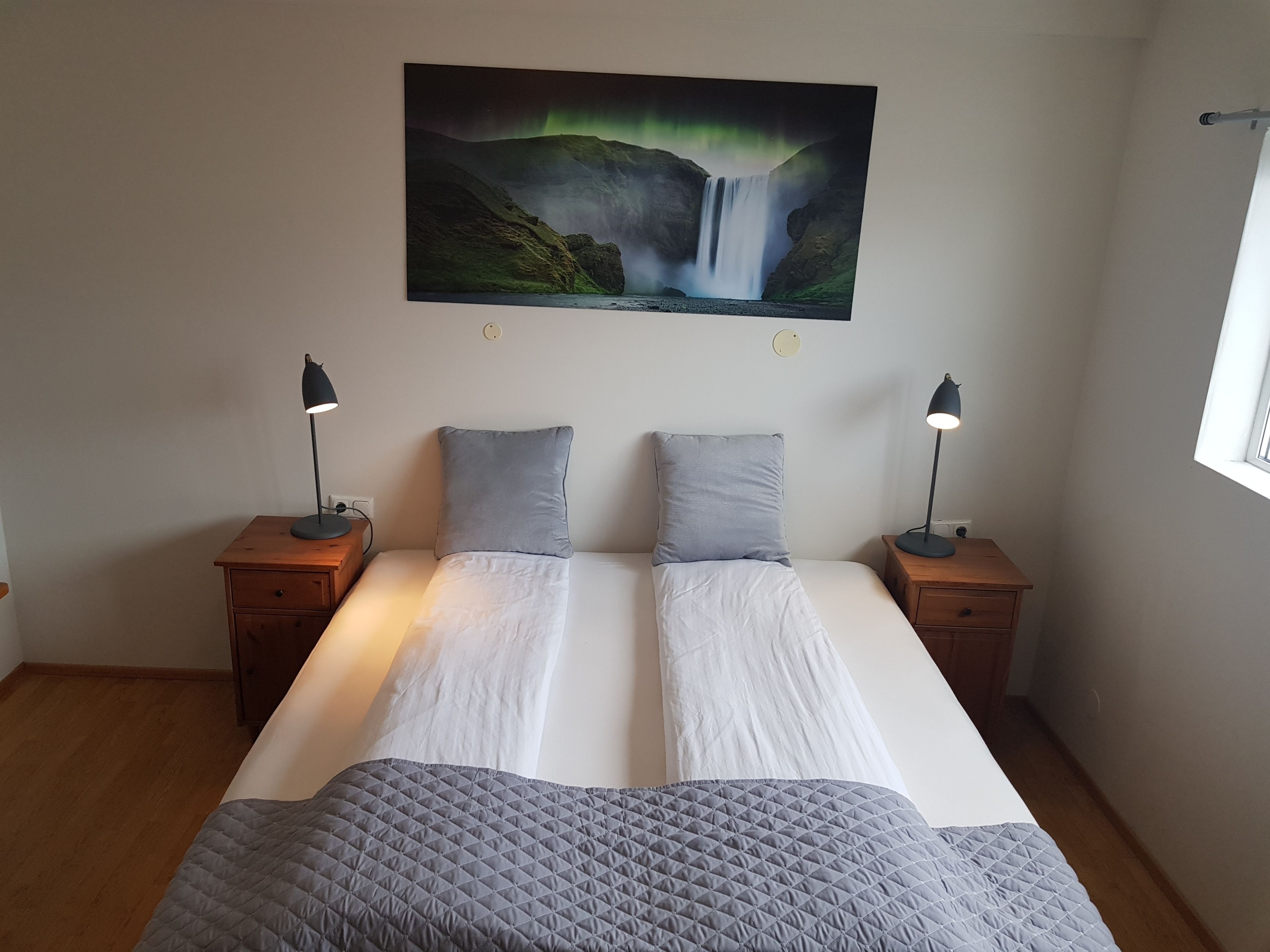 A double bed with a quilted gray blanket, two gray pillows, and two bedside lamps on wooden nightstands. A large painting of a waterfall and green lights is on the wall above the bed.