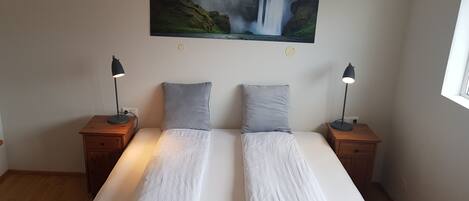 A double bed with a quilted gray blanket, two gray pillows, and two bedside lamps on wooden nightstands. A large painting of a waterfall and green lights is on the wall above the bed.