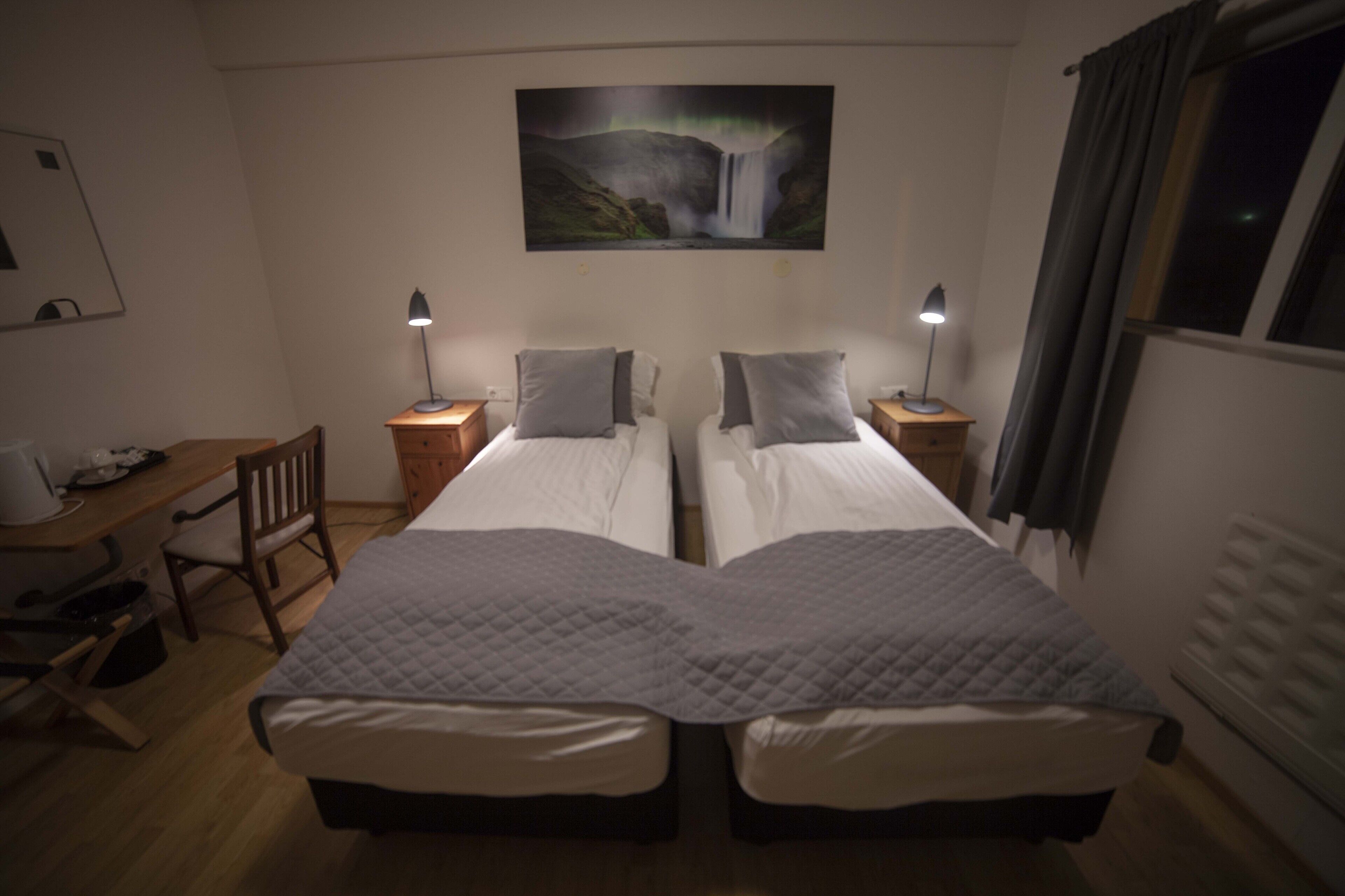 Una habitación con dos camas, un escritorio y una silla. Hay un cuadro en la pared y una ventana con cortinas.