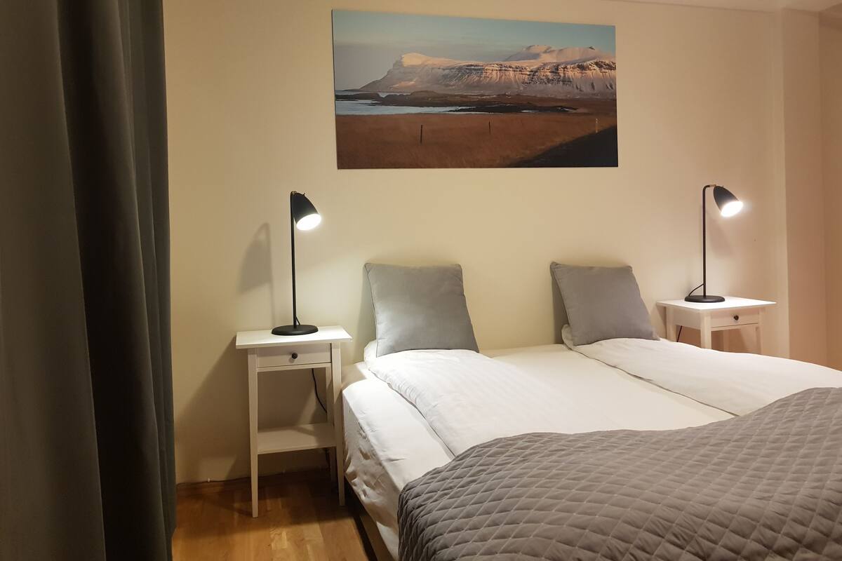 A hotel room with a bed, two bedside tables, a nightstand, a painting on the wall, and a curtain.