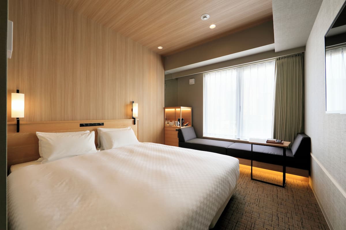 A hotel room with a large bed, a desk, and a chair. There is a window with curtains, a nightstand, and a lamp.