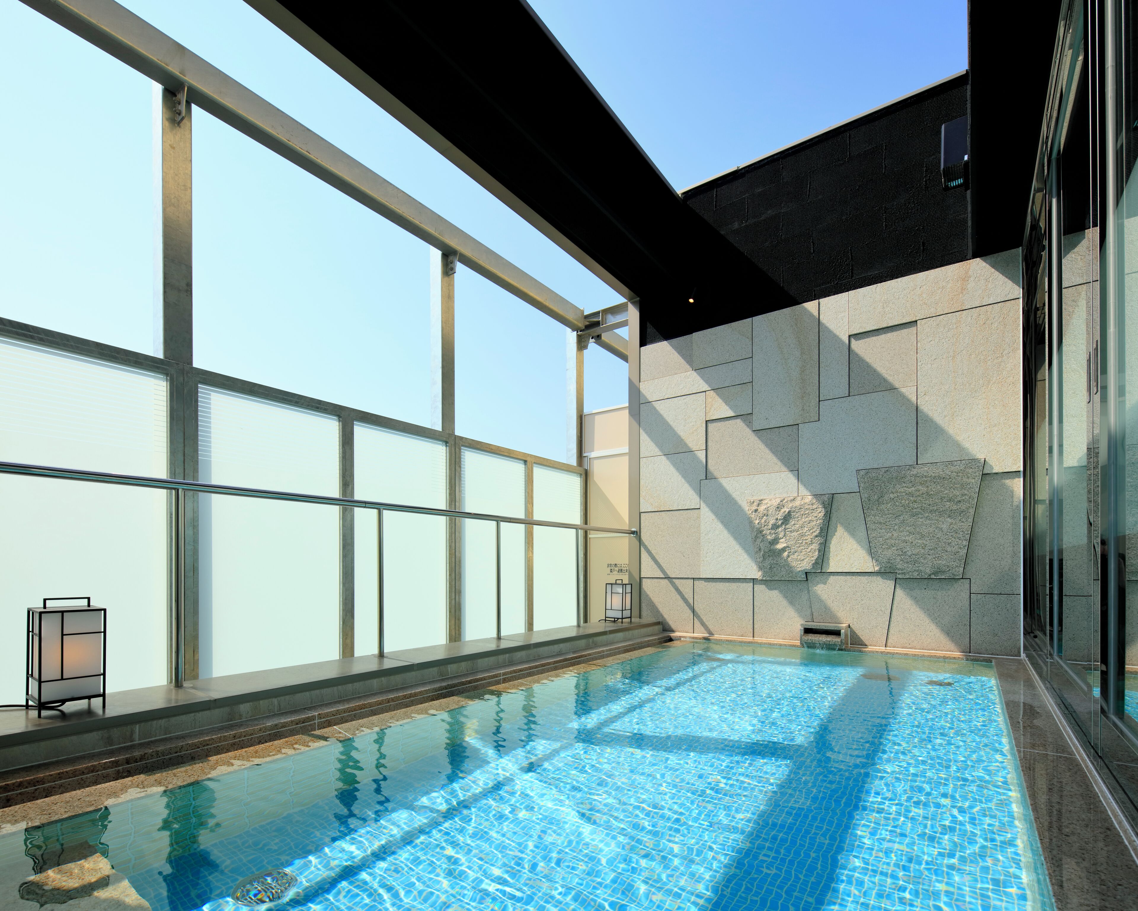 A swimming pool with clear blue water, large windows, and a stone wall.