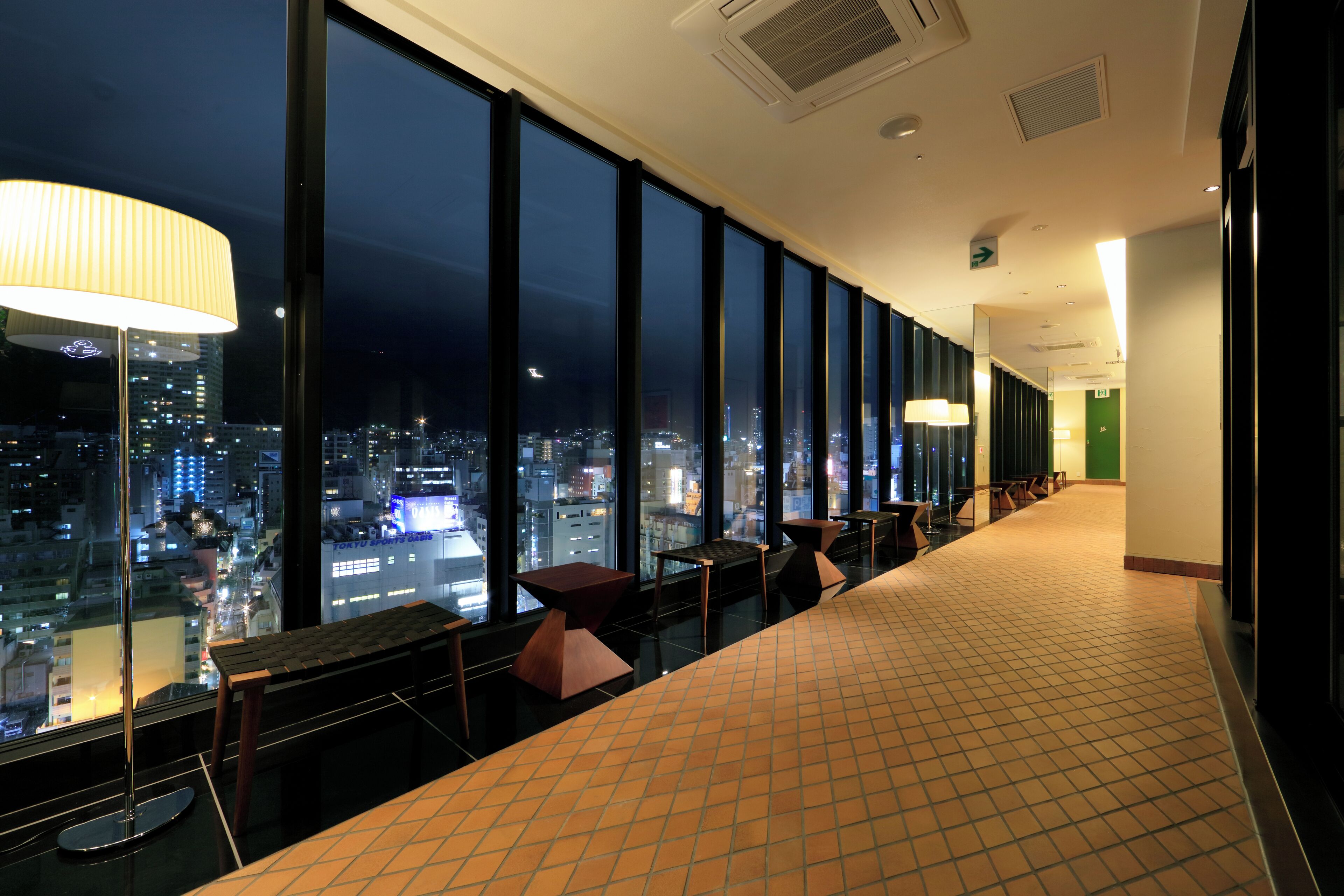 A hotel corridor with large windows offering a nighttime city view, a long reception desk, and a tiled floor.