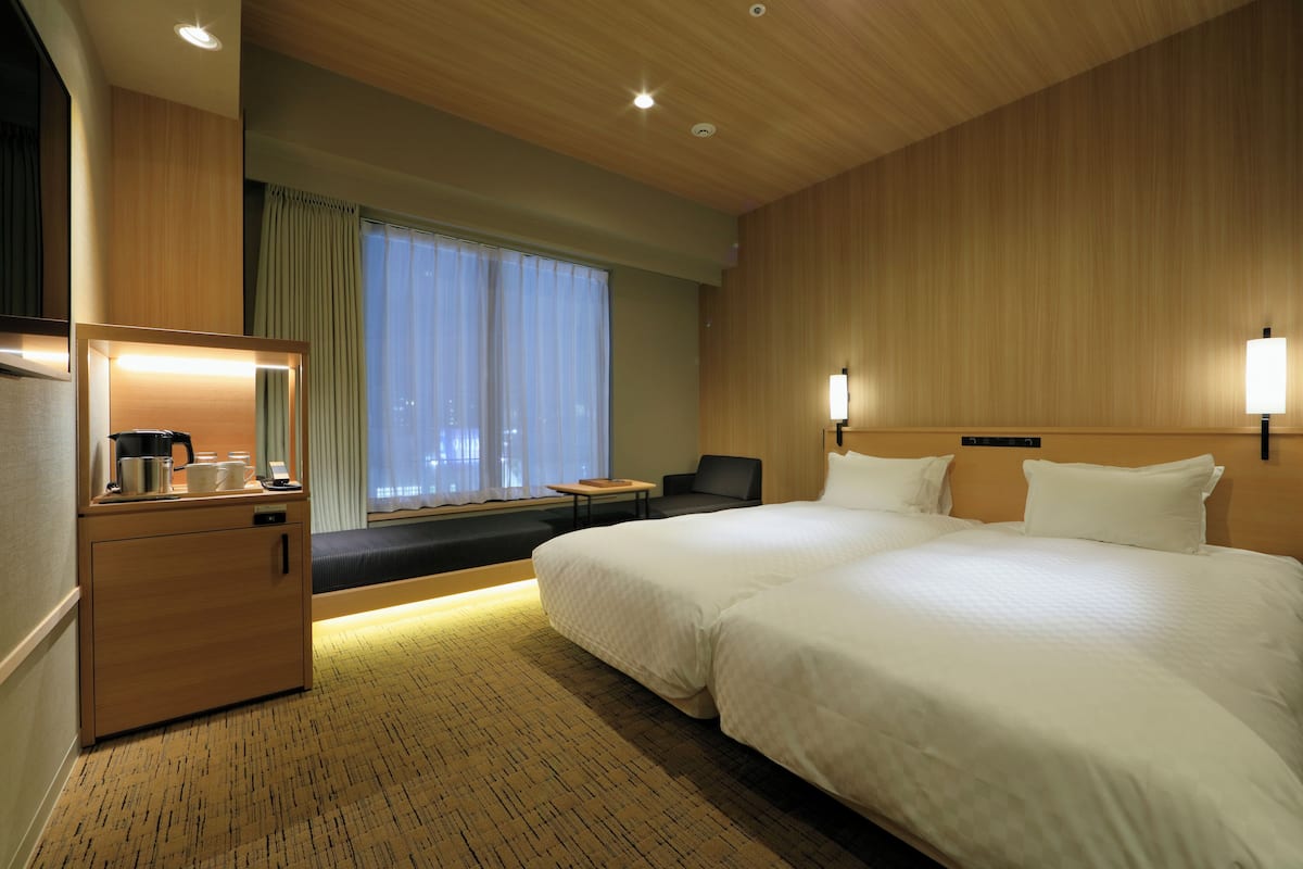 A hotel room with a large bed, a desk with a lamp, a chair, and a view of the city through large windows.