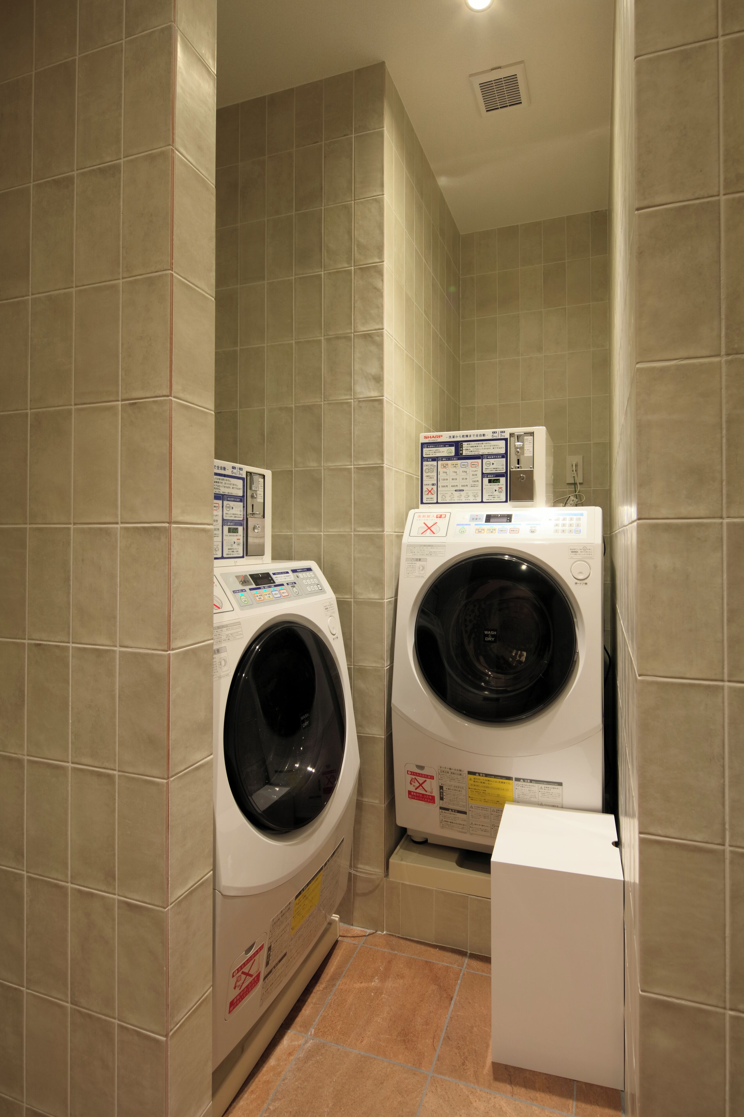 A compact laundry room with two front-loading washing machines and a dryer, enclosed by beige tiled walls.