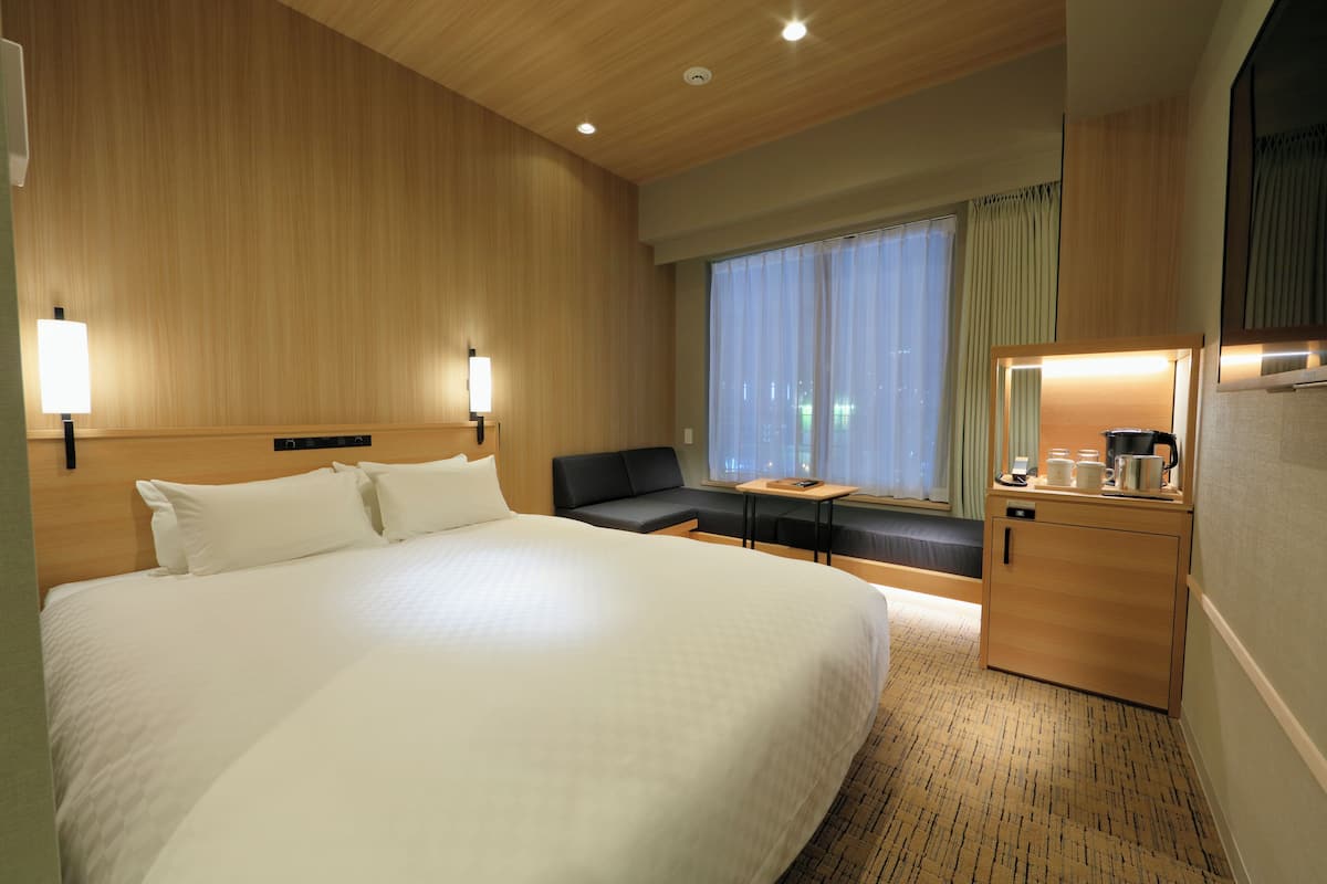 A hotel room with a large bed, a desk with a chair, a small cabinet, and a window with curtains.