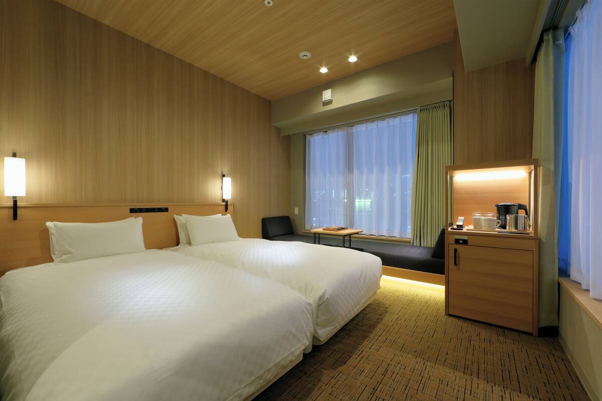A hotel room with two large beds, a small table, a desk with a lamp, and a view of the city through large windows.