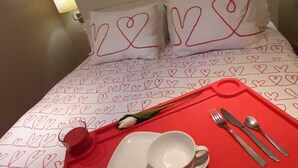 Habitación de hotel con una cama y una colcha estampada con motivos de corazón, una bandeja roja con una taza y cubiertos, y arte en la pared que representa monumentos famosos.